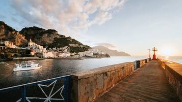 A coastal town with a wooden pier leading to a lighthouse, overlooking a calm sea and a mountainous backdrop.