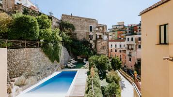 A pool area with a wooden deck, surrounded by stone walls and greenery, overlooking a narrow street with buildings.