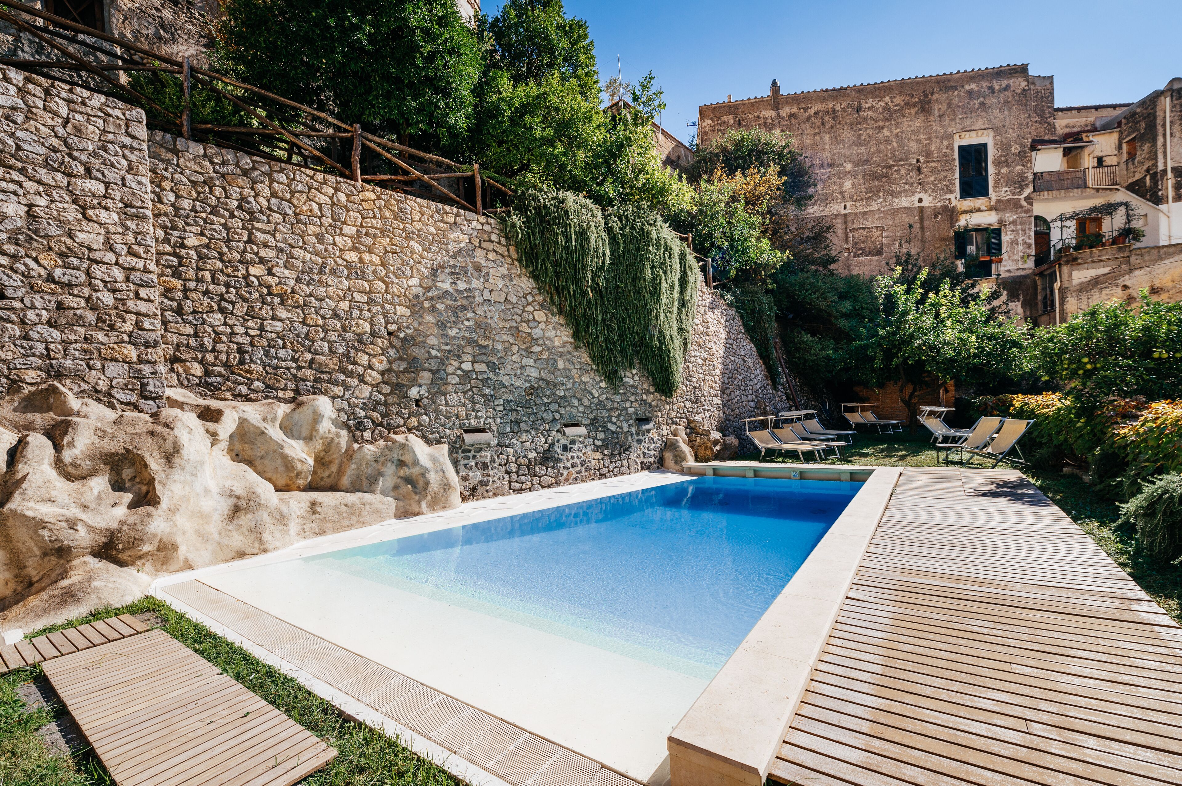 Ein Schwimmbad mit Holzterrasse, Steinmauer und Blick auf alte Gebäude.