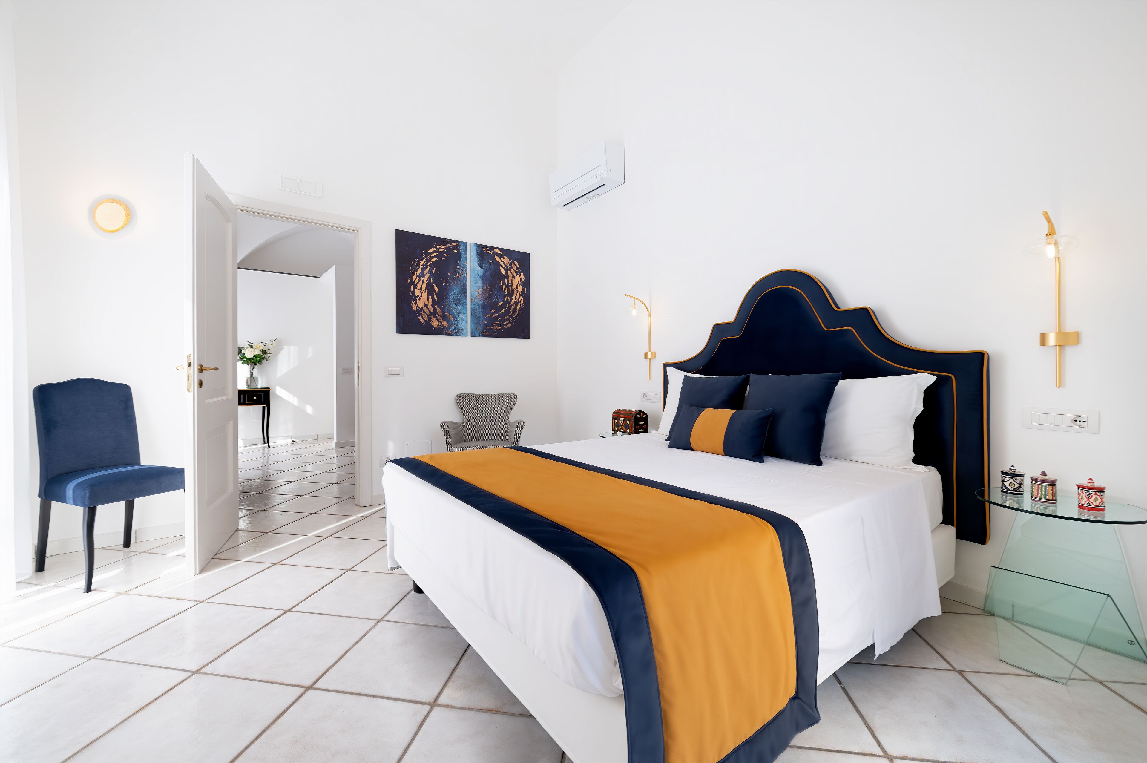 A modern hotel room with a large bed, a chair, a glass table, and a painting on the wall.