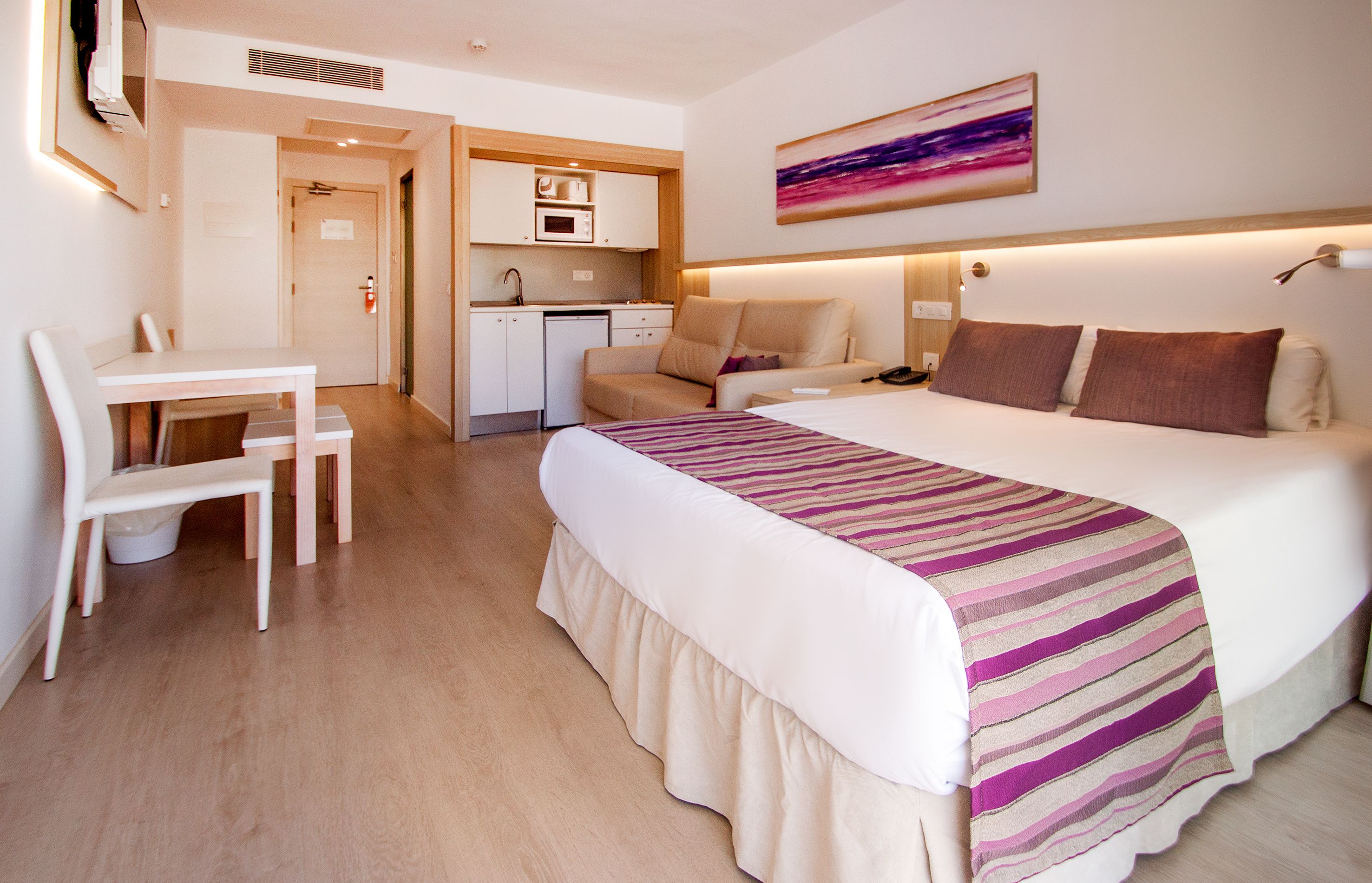 A hotel room with a bed, a desk, a chair, a kitchenette, and a painting on the wall.