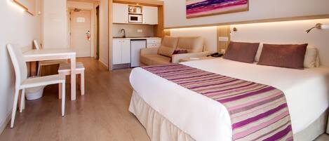 A hotel room with a bed, a desk, a chair, a kitchenette, and a painting on the wall.