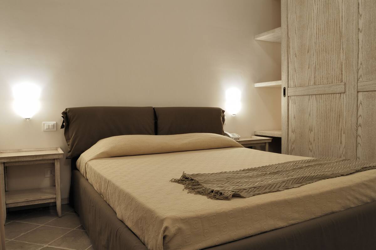 Un lit bien fait, recouvert d’une couvre-lit beige et agrémenté d’un plaid décoratif, est flanqué de deux tables de chevet sur lesquelles reposent des lampes. La pièce est également dotée d’une armoire en bois et d’un mur sobre.
