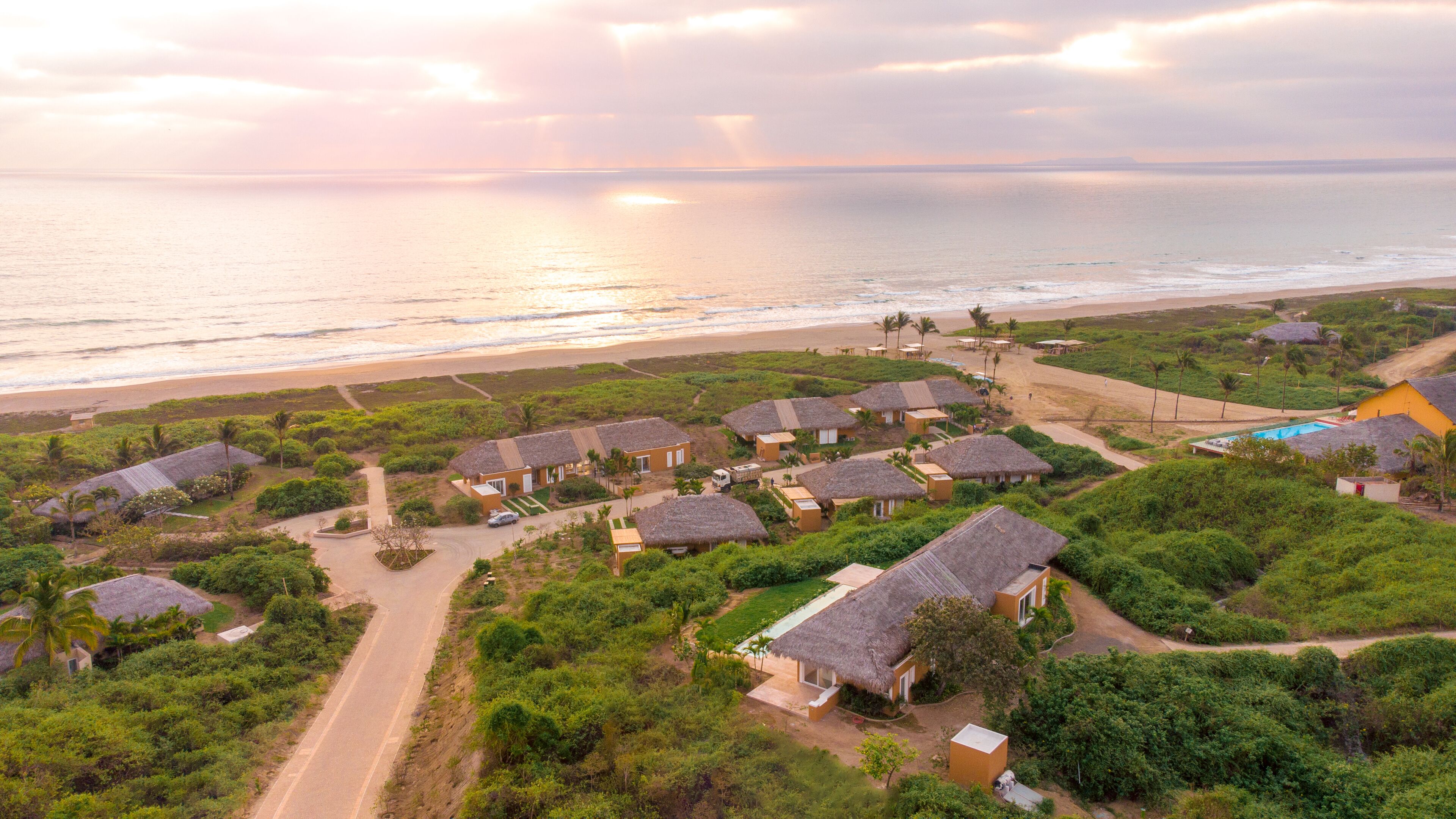 An aerial view of a coastal resort with thatched-roof cottages, a swimming pool, and a beach.