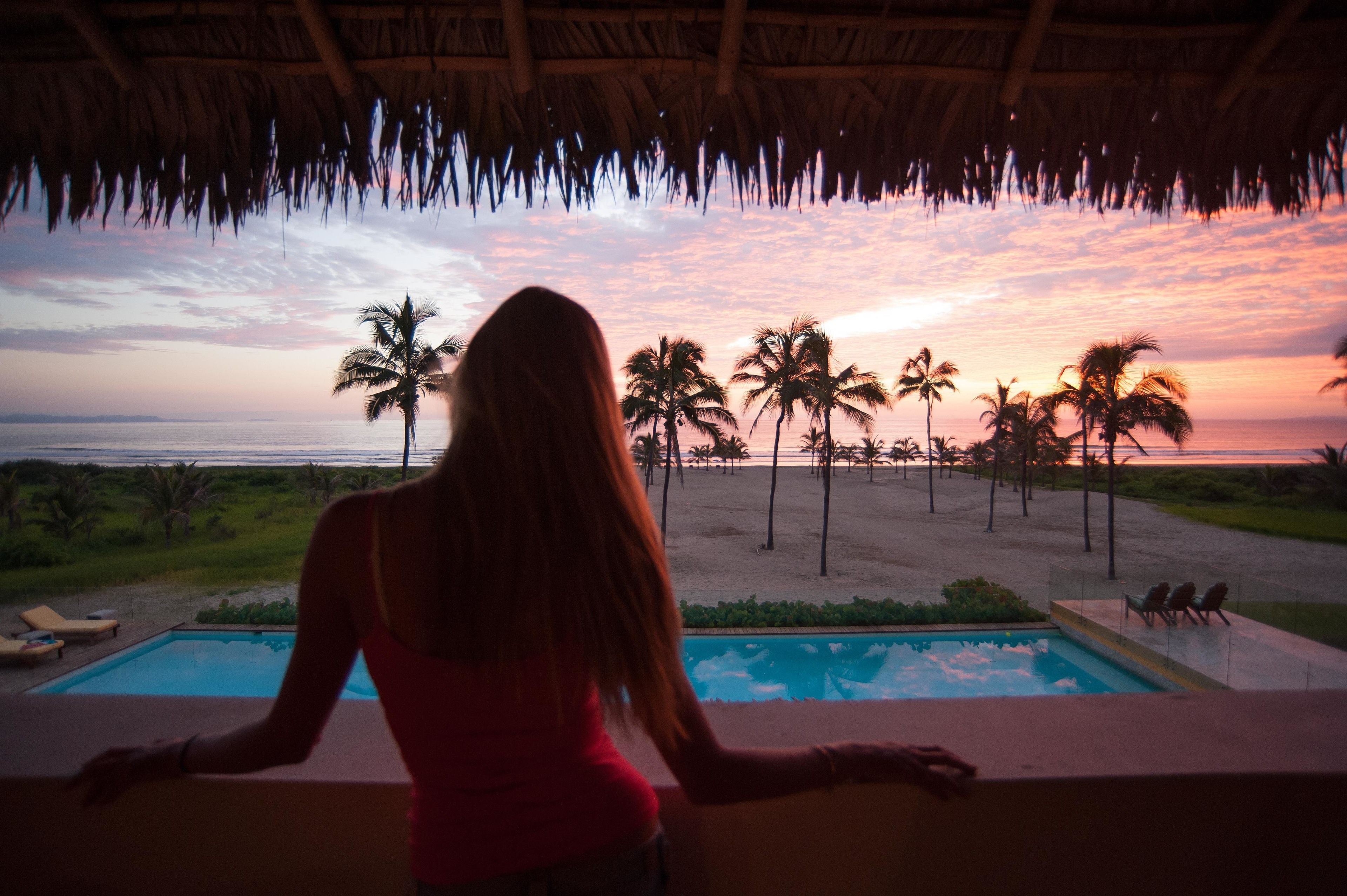 A person overlooking a pool with a view of palm trees and the ocean at sunset.