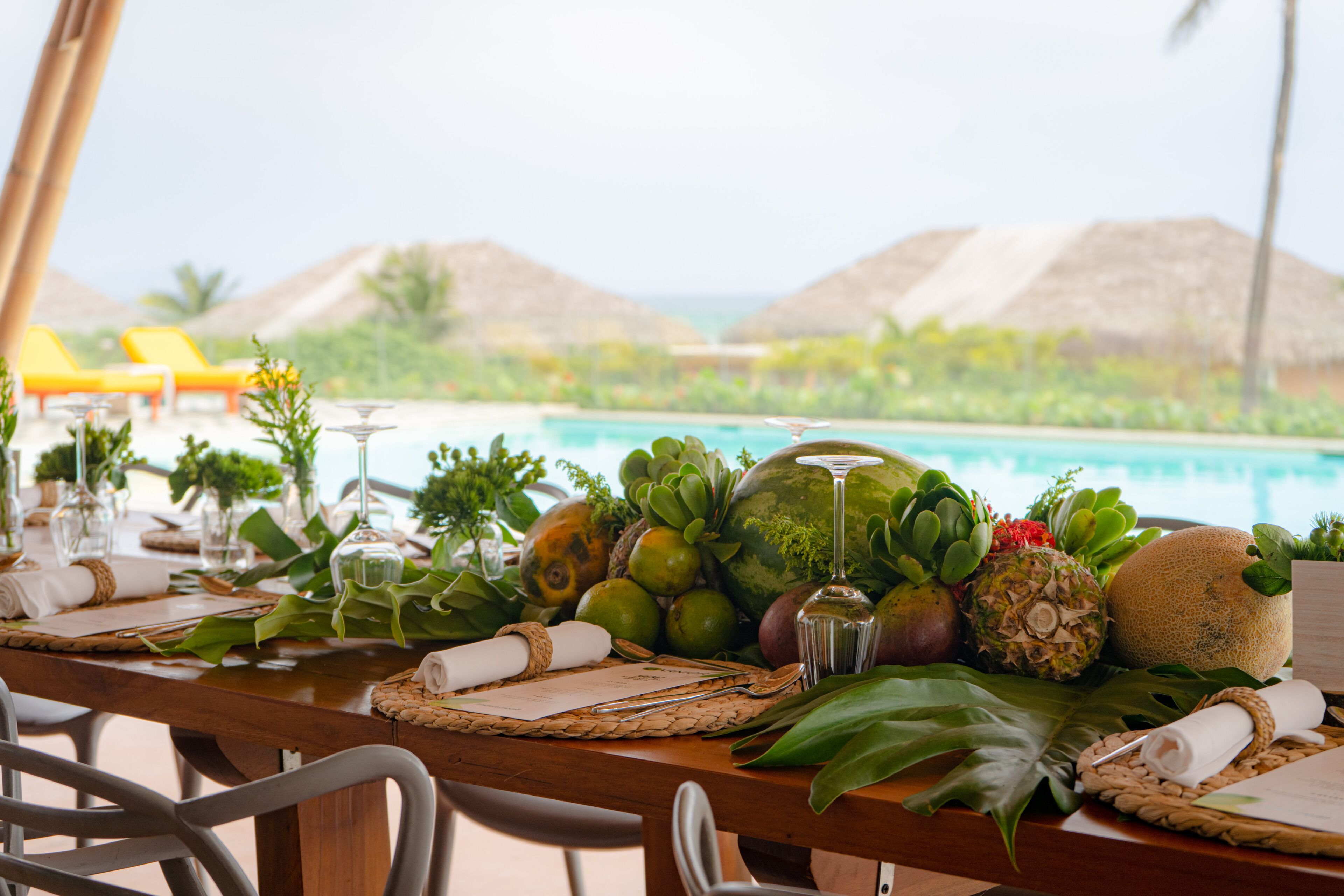 Mesa dispuesta para una comida con frutas tropicales, una copa de vino y vistas a una piscina y palmeras.