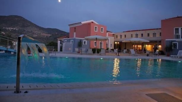 A swimming pool with a waterfall feature, surrounded by buildings with a Mediterranean architectural style, under a twilight sky. - Theofilos Superior Resort Hotel & Spa (Lesvos)