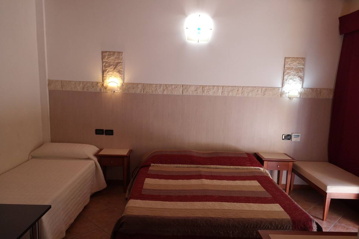 A hotel room with two single beds, a large bed with a striped blanket, a small table, a nightstand, and a wall-mounted light fixture.
