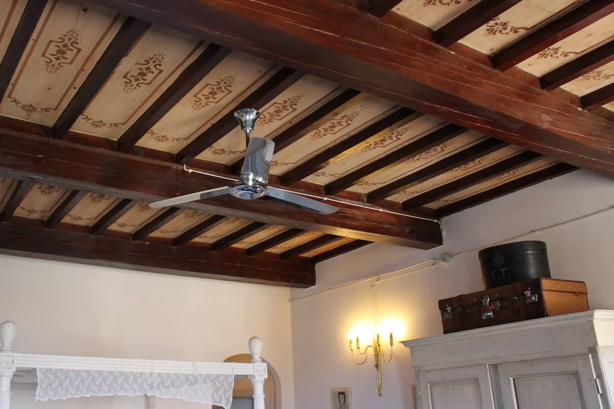A room with a wooden ceiling, a ceiling fan, a white table with a lace tablecloth, a wall-mounted light fixture, and a fireplace with a trunk above it.