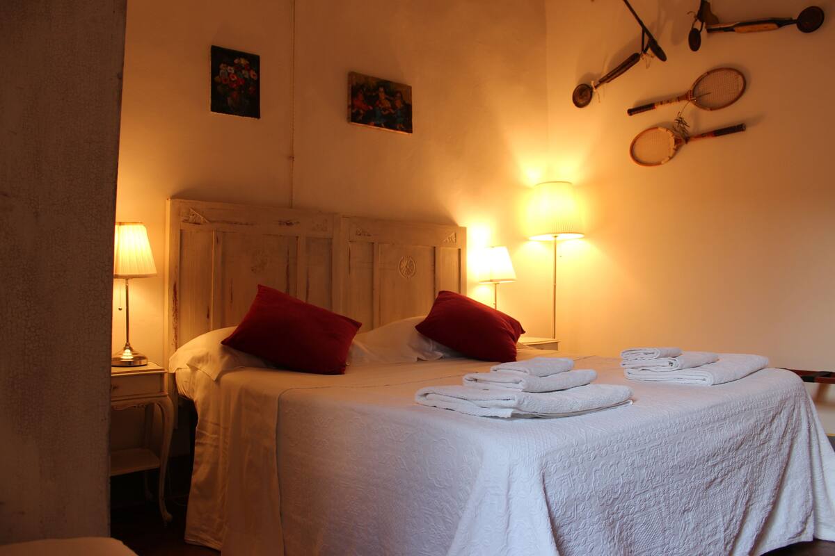 A neatly made bed with red pillows, a bedside table with a lamp, and a wall with sports equipment.