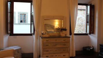 A room with a dresser, a mirror, a window with curtains, a small table, and a view of the outside.