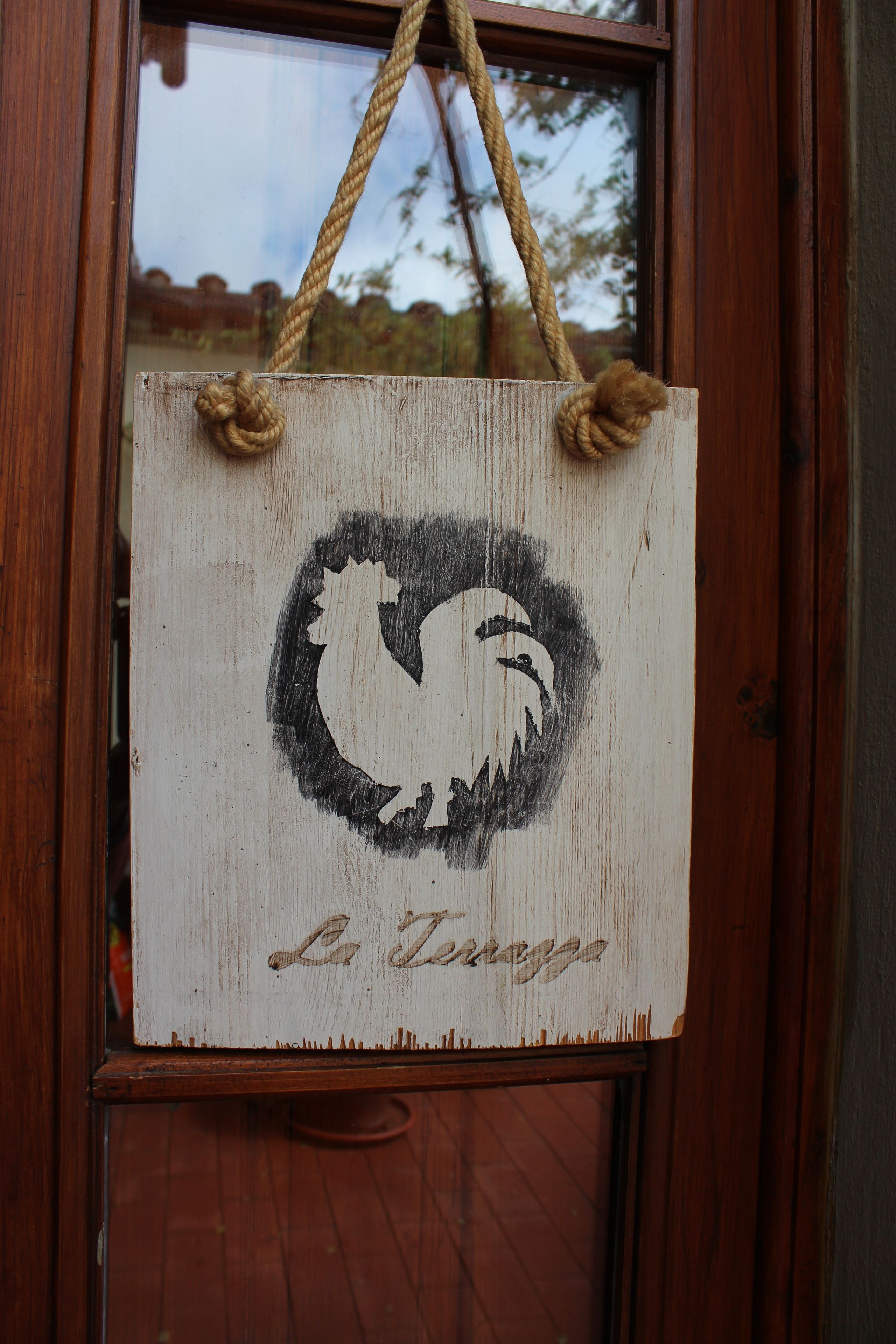 A wooden sign with a rooster logo and the text "La Terme" hanging on a window with a rope handle.