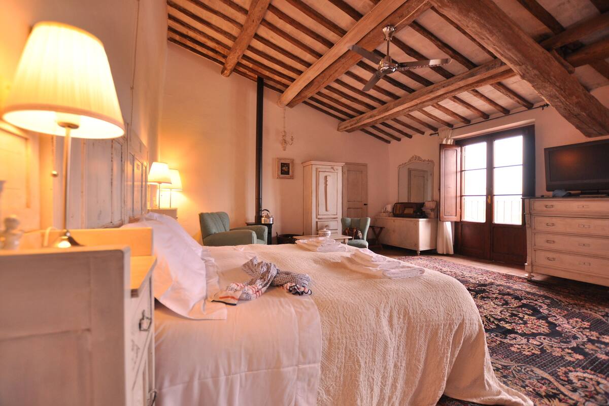 A spacious bedroom with a large bed, wooden beams on the ceiling, a television, and a window with natural light.