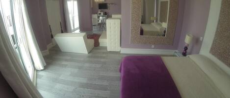A bedroom with a large bed, a dresser, a vanity with a mirror, and a kitchenette in the background.