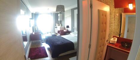 A hotel room with a bed, a desk, a chair, a small table, and a bathroom with a mirror and sink.