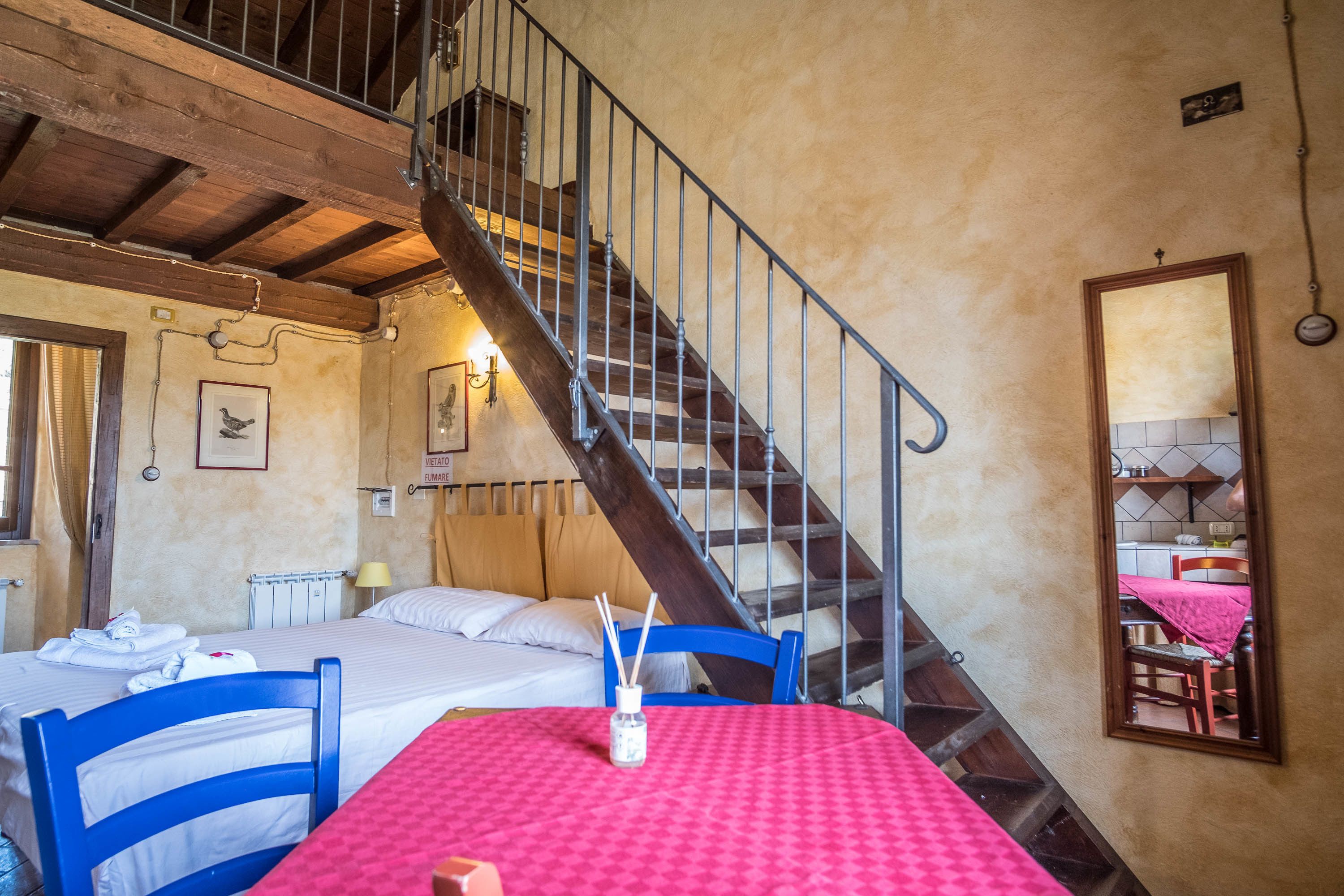 Una habitación con una cama, una mesa con mantel rojo, una silla azul, una escalera de madera y un espejo en la pared.