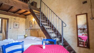 Una habitación con una cama, una mesa con mantel rojo, una silla azul, una escalera de madera y un espejo en la pared.