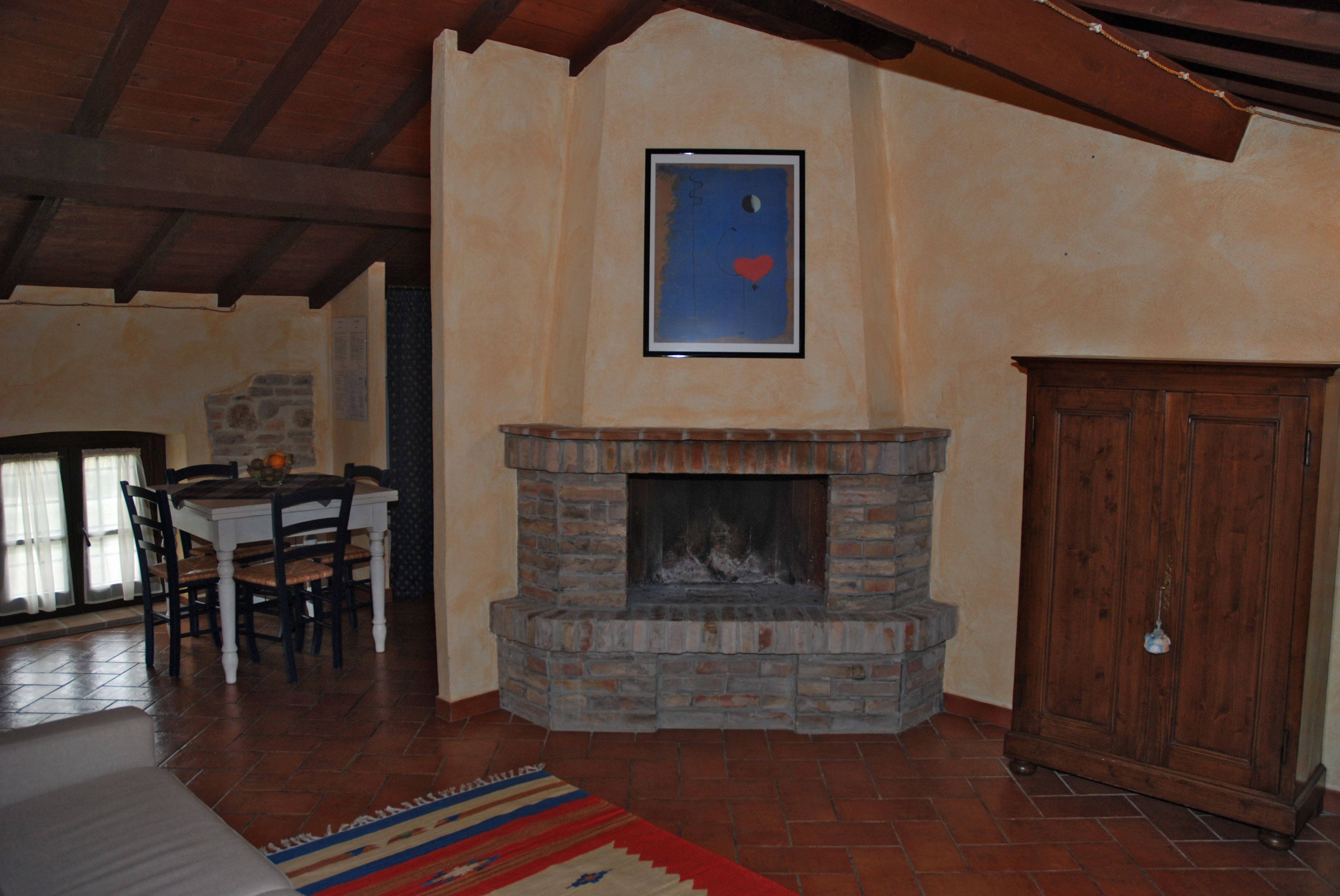 A living room with a fireplace, wooden ceiling beams, a dining table with chairs, and a patterned rug.