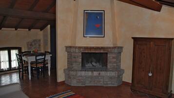 A living room with a fireplace, wooden ceiling beams, a dining table with chairs, and a patterned rug.