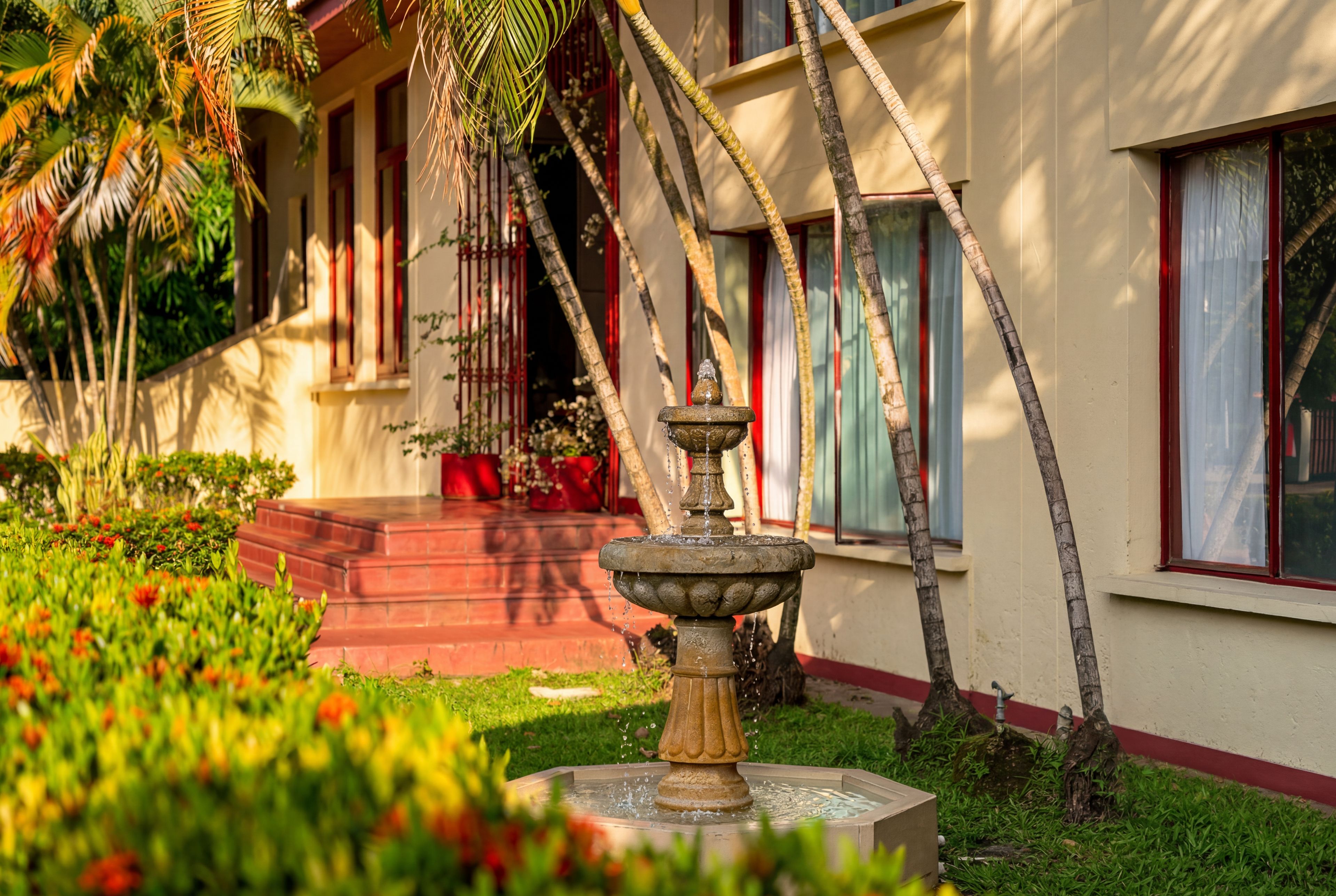 Un jardín con una fuente, palmeras y un edificio con detalles en rojo.