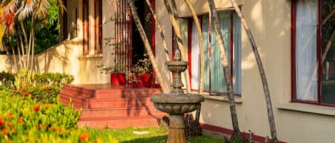 Un jardín con una fuente, palmeras y un edificio con detalles en rojo.