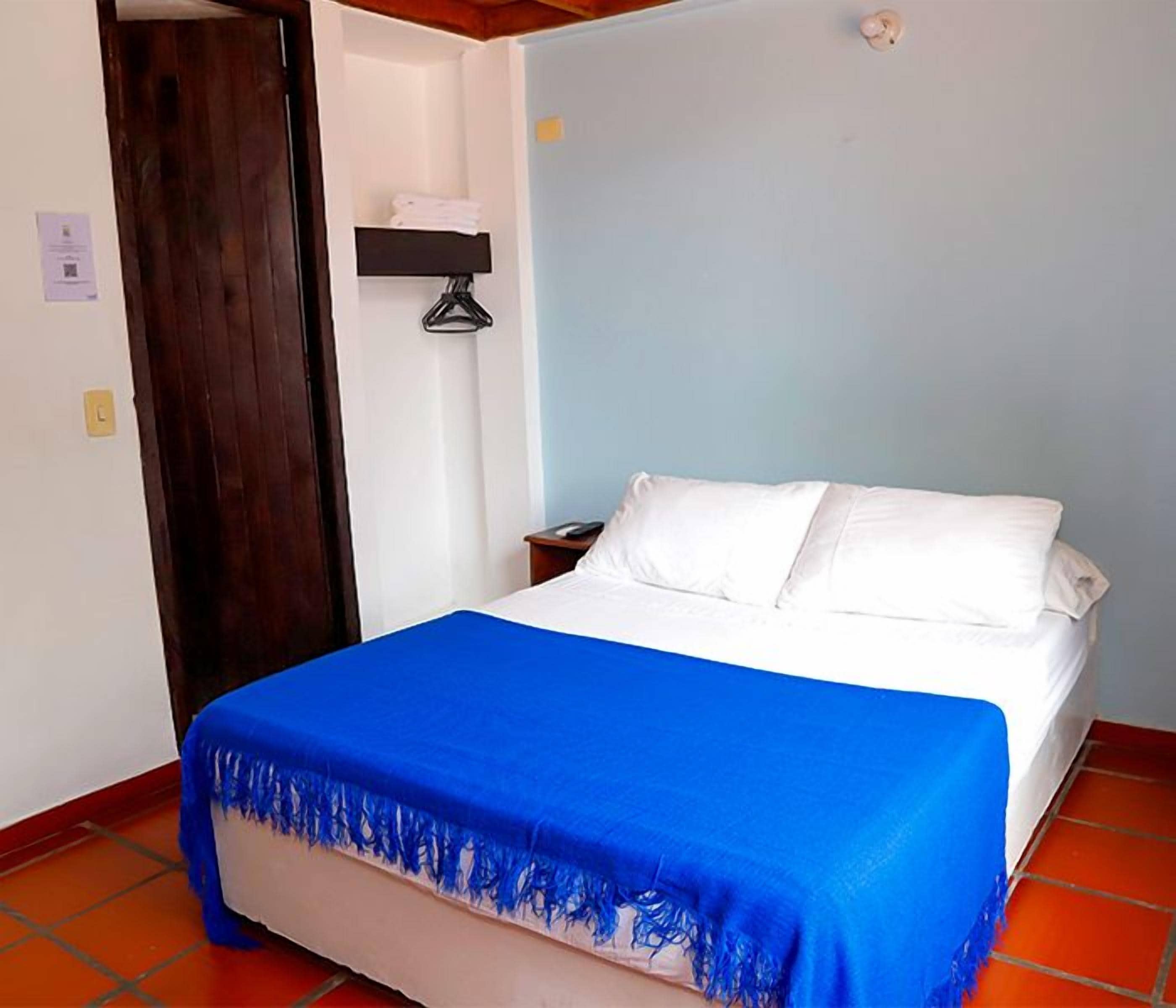 Un dormitorio con una cama, una manta azul, una puerta de madera y un estante fijado a la pared.