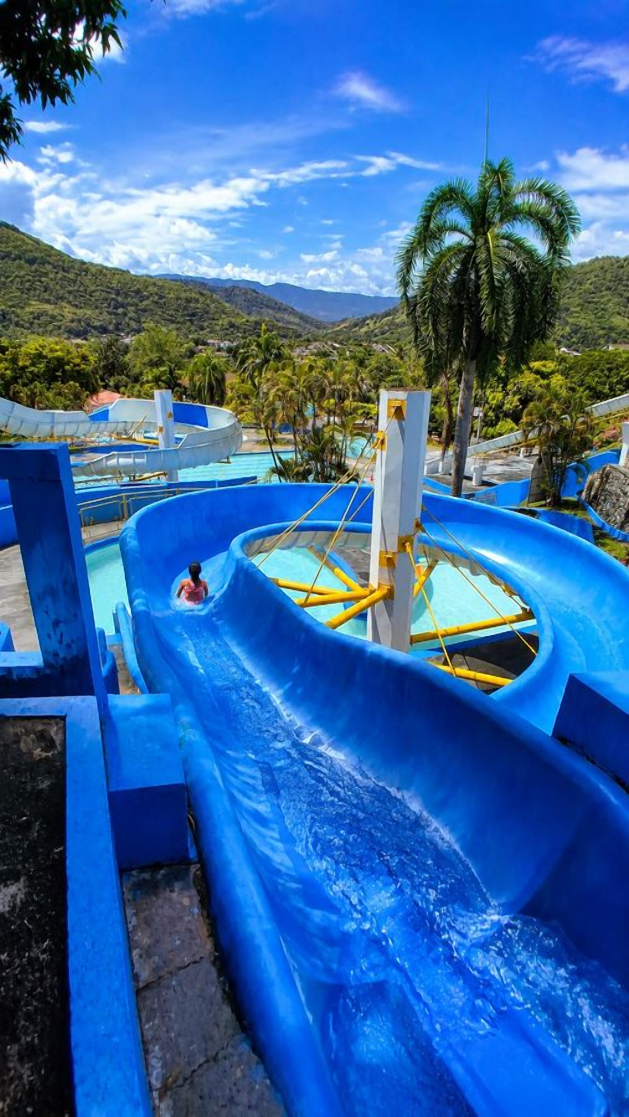Uno scivolo acquatico con una persona in fondo, circondato da una piscina e palme, con un paesaggio montuoso sullo sfondo.