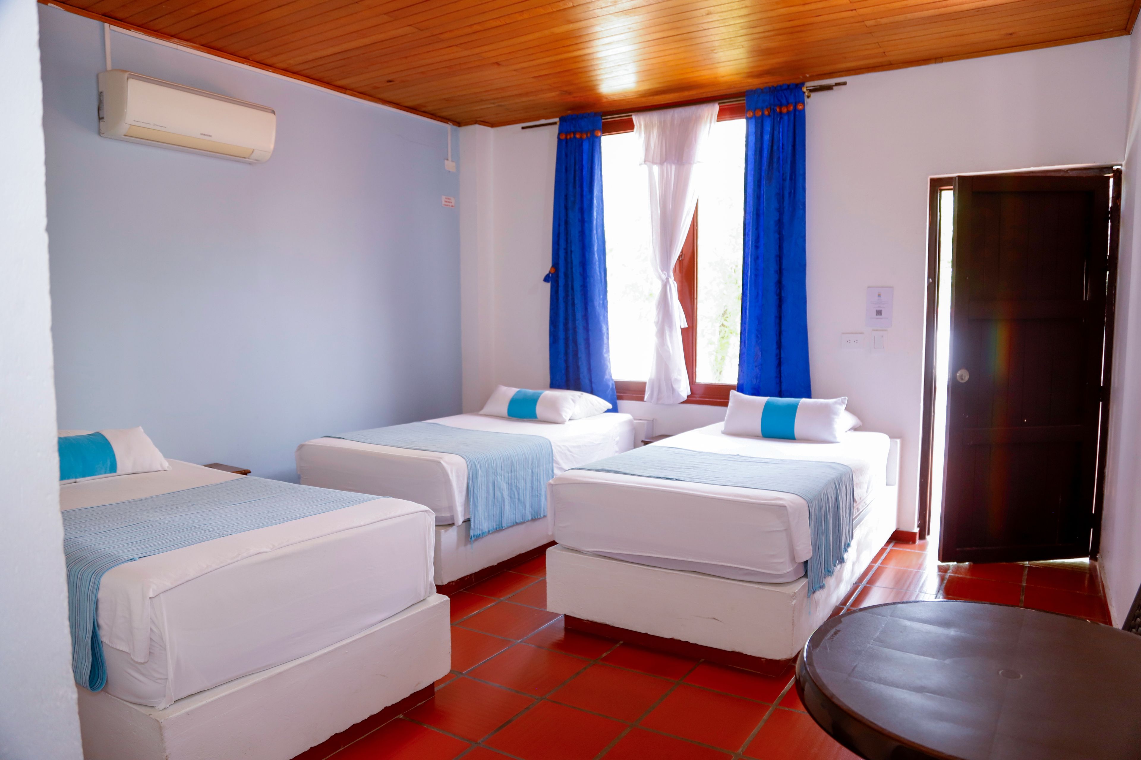 Una habitación de hotel con dos camas individuales, una mesita pequeña, un ventilador de techo y una ventana con cortinas.