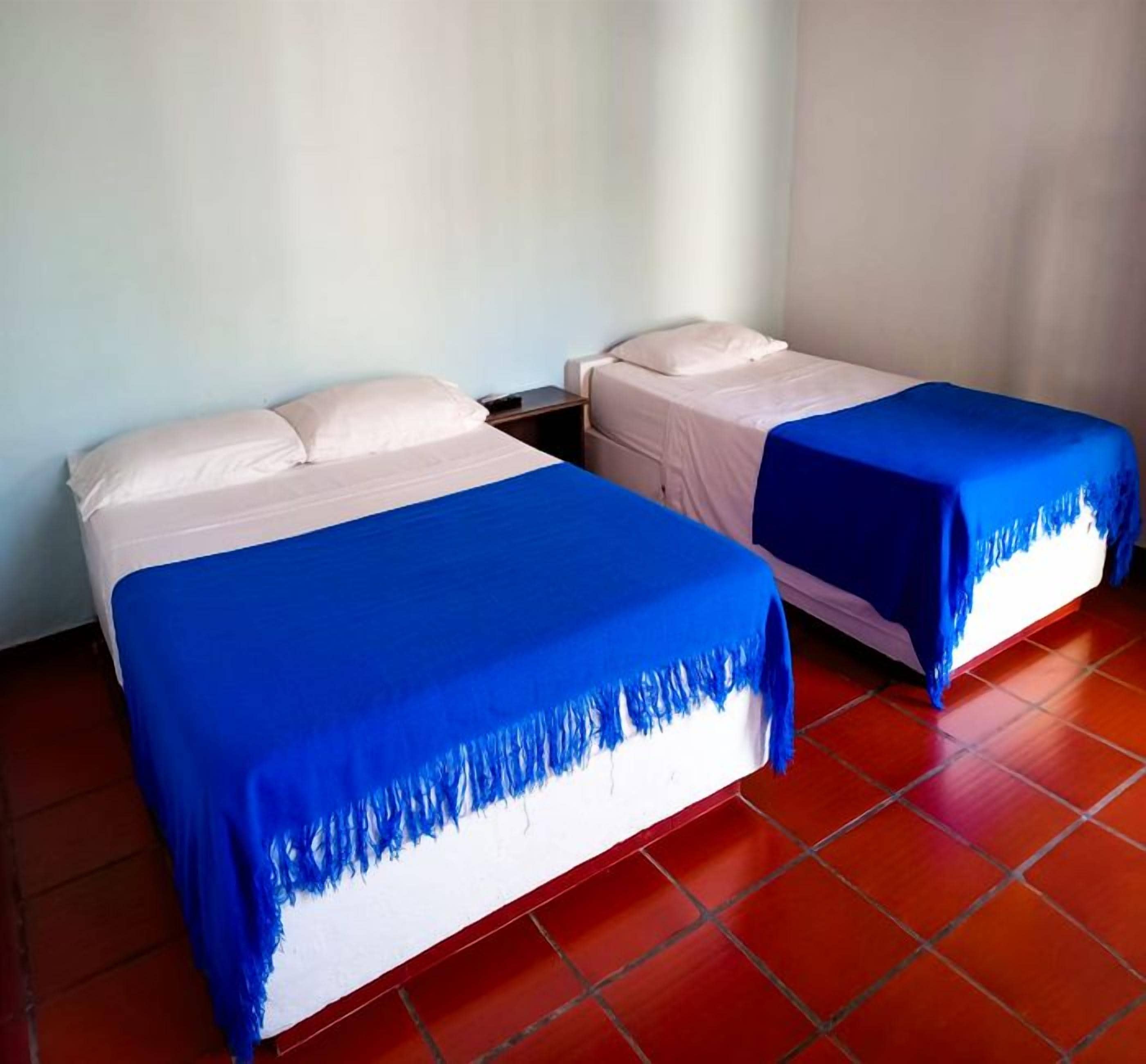 Duas camas de solteiro com lençóis brancos e colchas azuis em um quarto com piso de ladrilho vermelho.