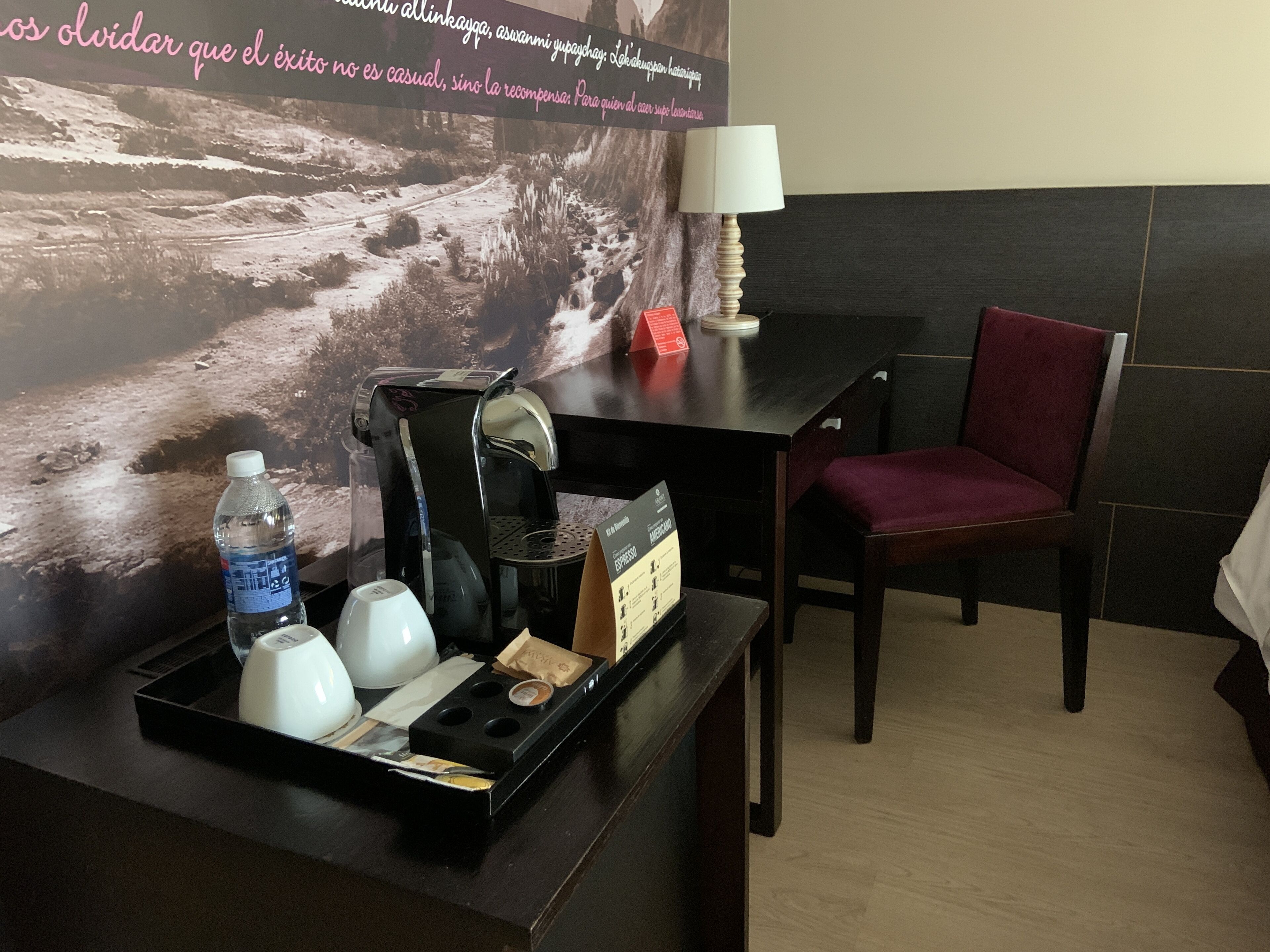 A hotel room with a desk, a chair, a coffee maker, a water bottle, and a tray with cups and a bag.