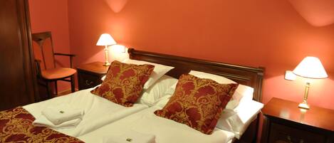 Una cama bien hecha con sábanas blancas y almohadones estampados en rojo, dos mesitas de noche con lámparas, y una silla en una habitación con paredes rojas.