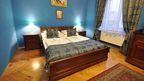 A bedroom with a wooden bed, two bedside tables with lamps, a blue wall, a framed picture, and a window with curtains. - Grand Hotel Praha (Jicin)