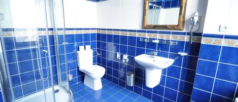 Un baño con azulejos azules, mampara de ducha de cristal, inodoro, lavabo y espejo con marco decorativo.