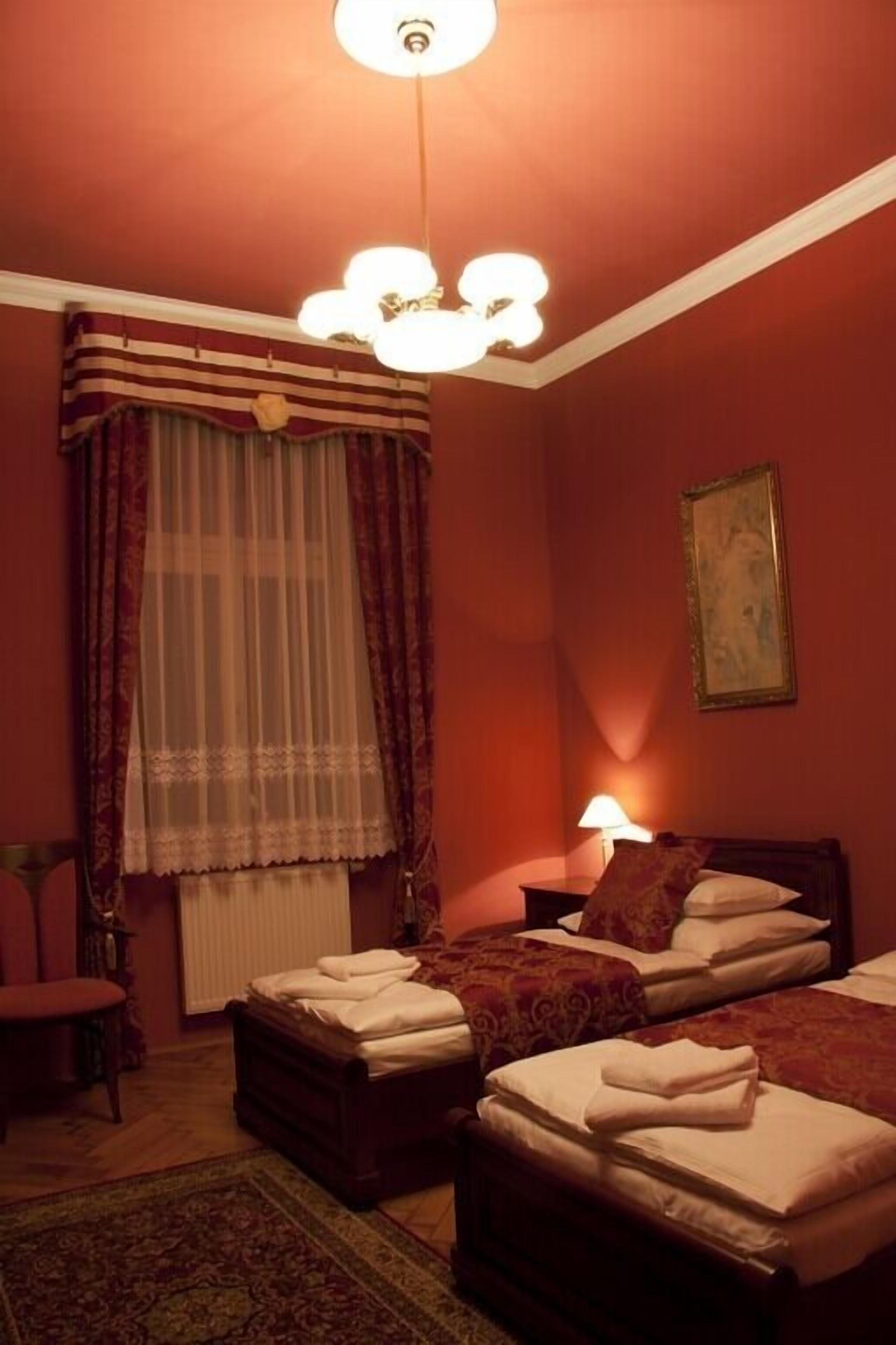 A hotel room with two beds, a wooden headboard, a bedside table with a lamp, a window with curtains, and a framed picture on the wall.