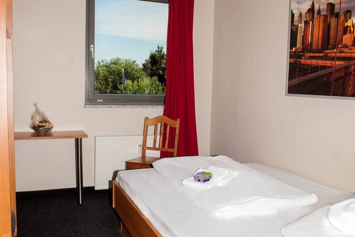 Ein Hotelzimmer mit Bett, Schreibtisch, Stuhl, Fenster mit rotem Vorhang und Blick auf die Stadt.