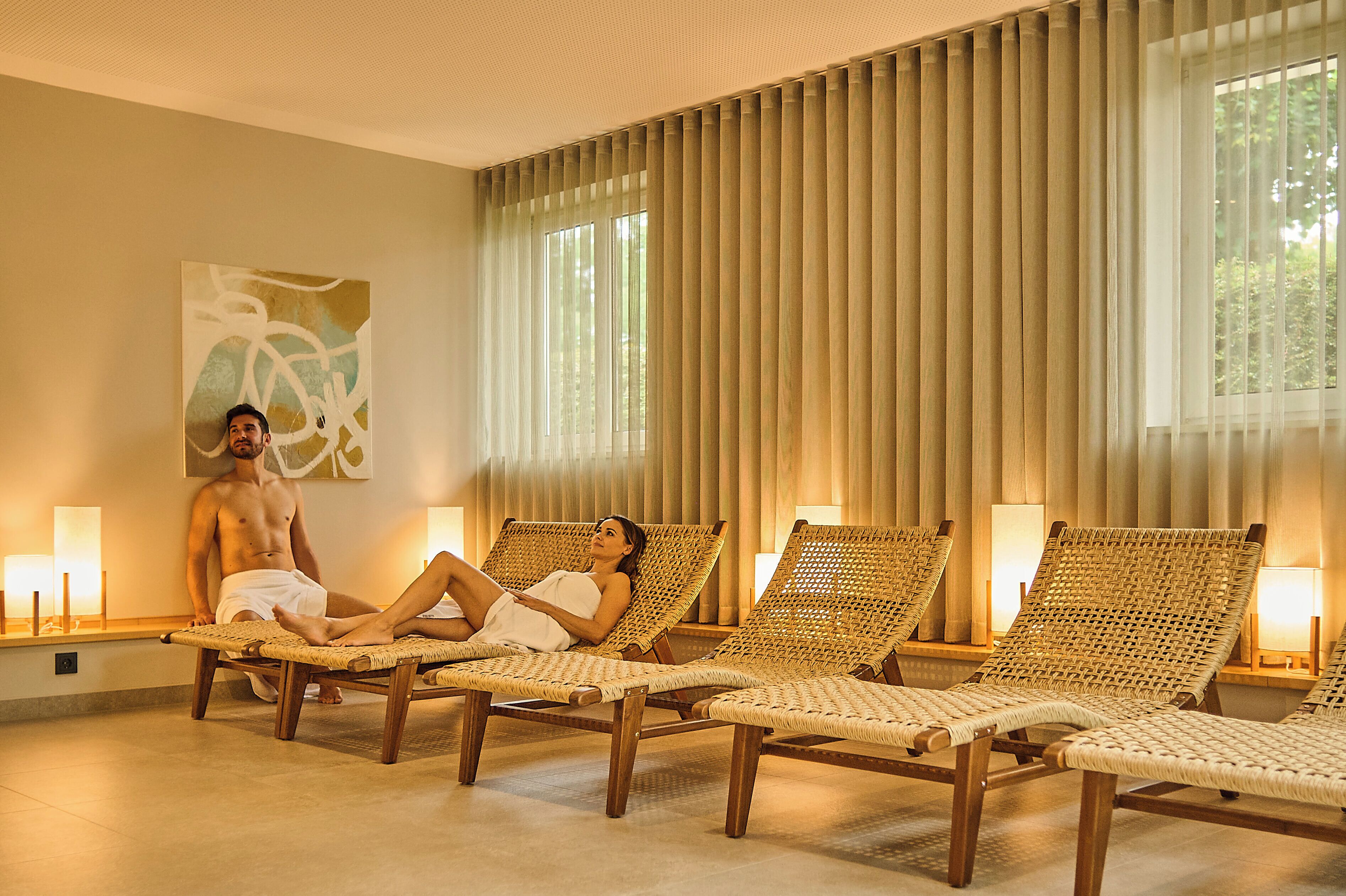 Une pièce comprenant trois fauteuils de relaxation en osier, un grand tableau abstrait et une vue sur les arbres par les fenêtres.