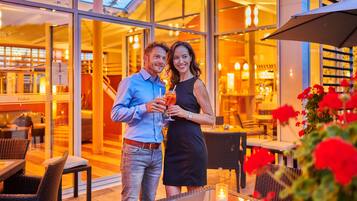 Una pareja parada en una mesa al aire libre con bebidas, con vista a un restaurante con puertas de cristal y muebles de mimbre.