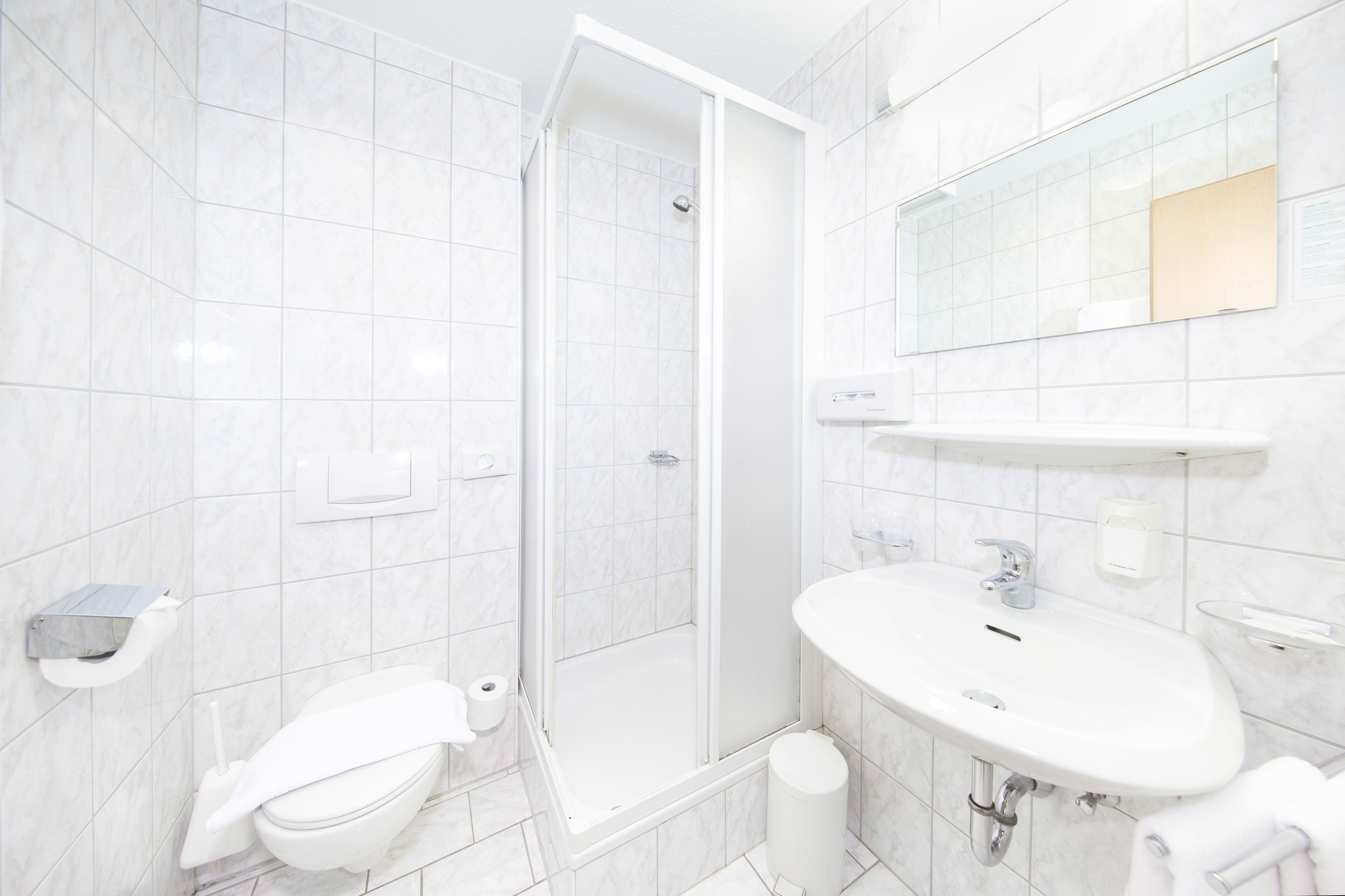 Ein modernes Badezimmer mit weißen Fliesen, einer Duschkabine, einem Waschbecken und einer Toilette.