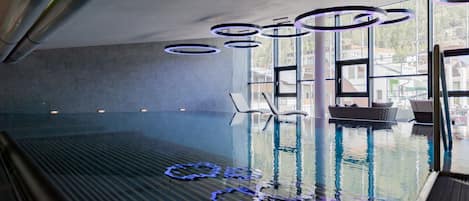 En inomhuspool med ett glasgolv, solstolar och modern belysning.