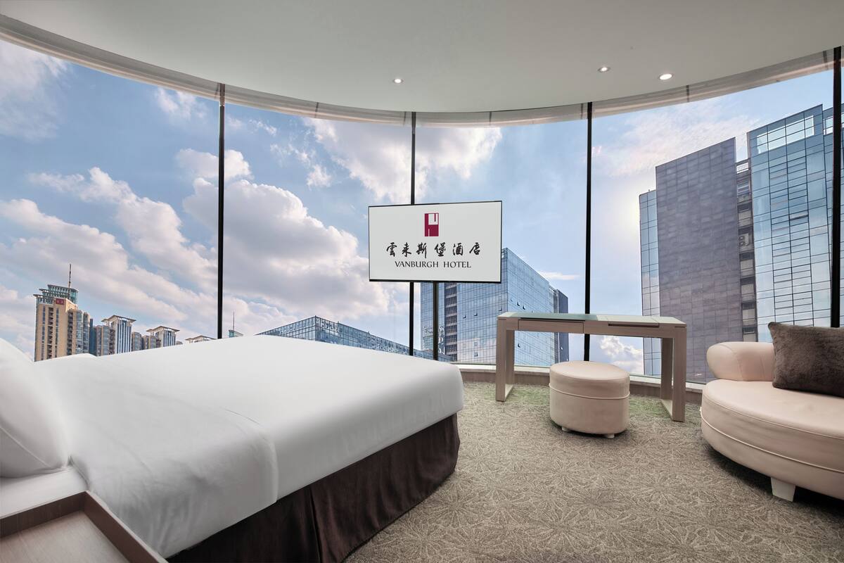 A modern hotel room with a large bed, a small table, and a sofa. A sign with the hotel name is visible.