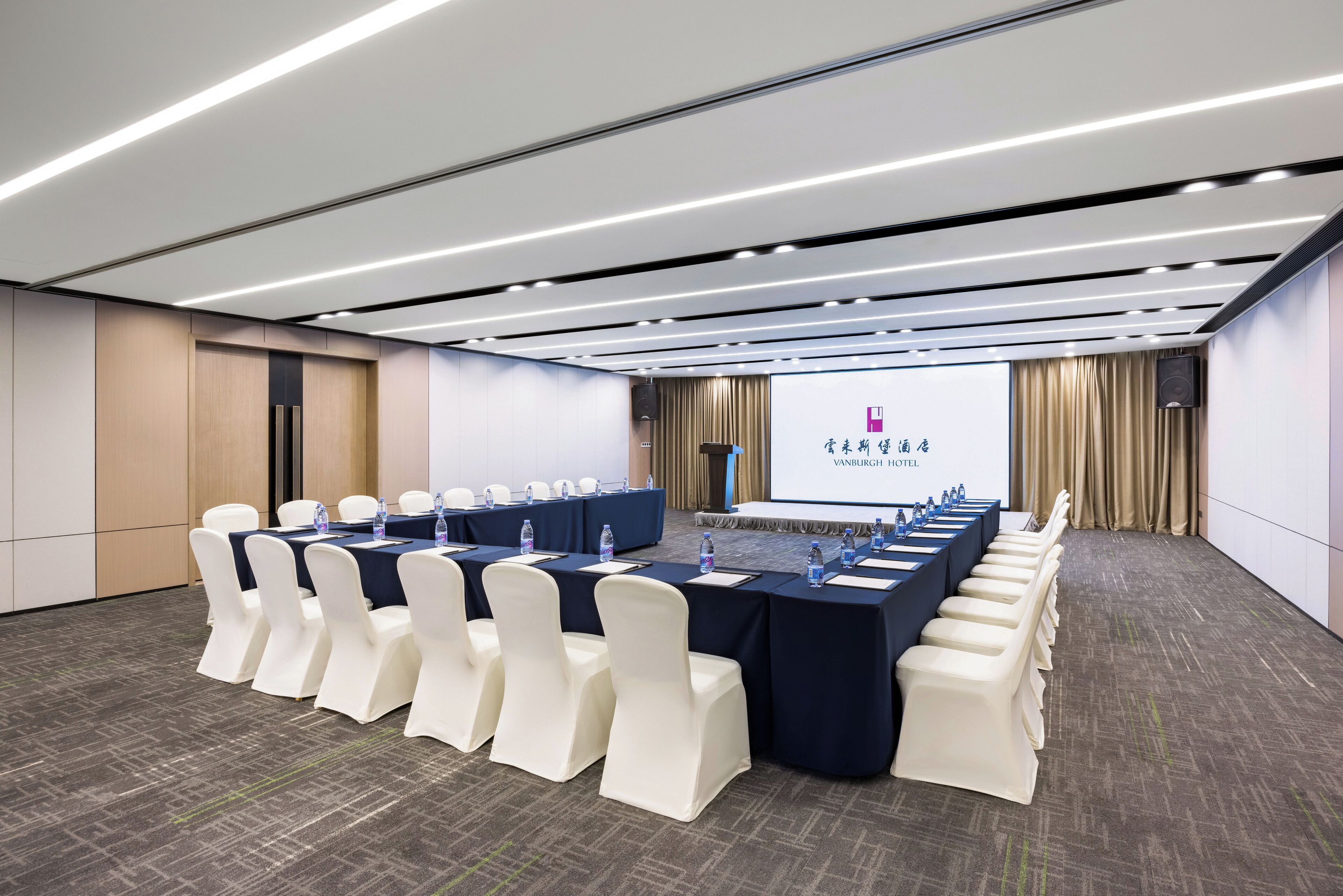 Un salón de conferencias con una mesa larga, sillas blancas y una pantalla mostrando una presentación.