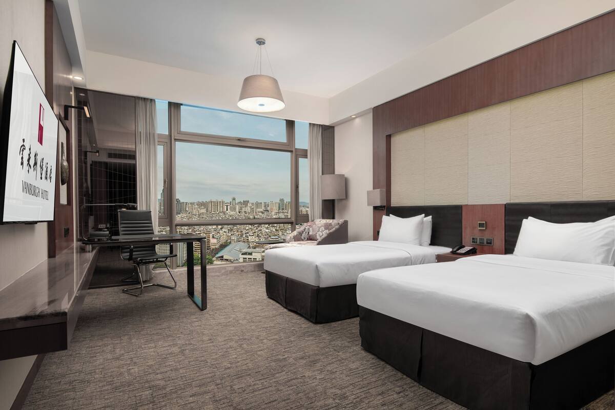 A hotel room with two beds, a desk, and a large window offering a city view.