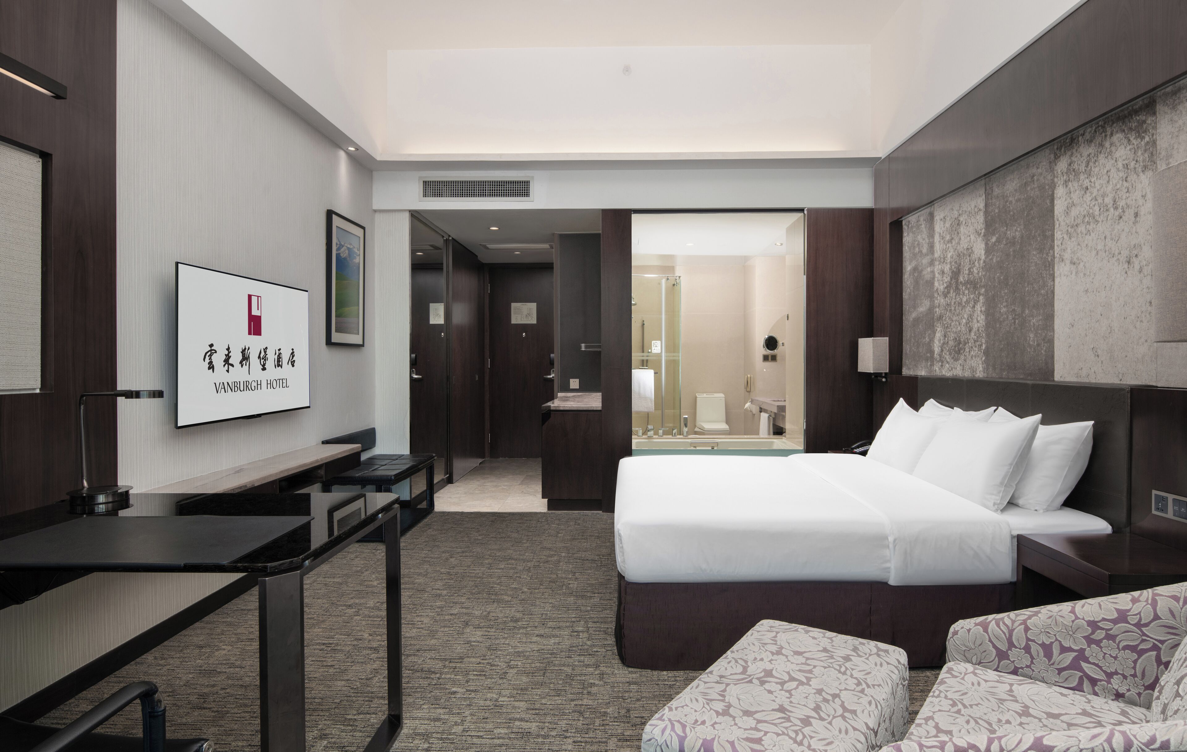 A hotel room with a bed, a desk, a chair, and a sofa. There is a bathroom visible through an open door.