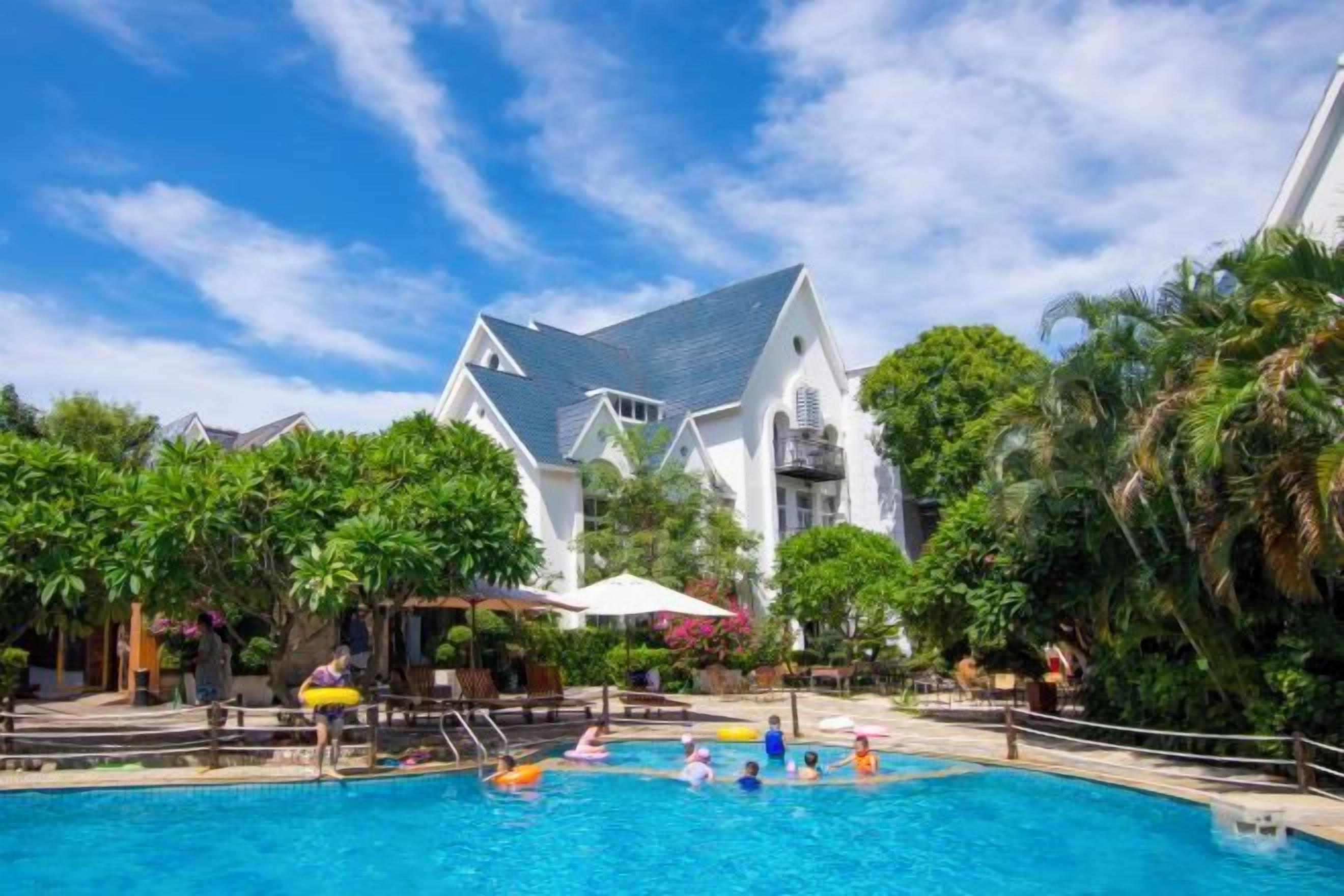 A swimming pool with people, surrounded by lush greenery and a large white building with a steep roof.