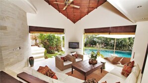 A spacious living room with a large white sofa, a wooden coffee table, and a television. The room has large windows with a view of a swimming pool and lush greenery. - Villa Sahaja (Kerobokan)