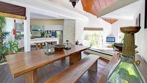 A spacious kitchen with a long wooden table, a glass-top dining table, and a modern kitchen area with a TV and plants. - Villa Sahaja (Kerobokan)
