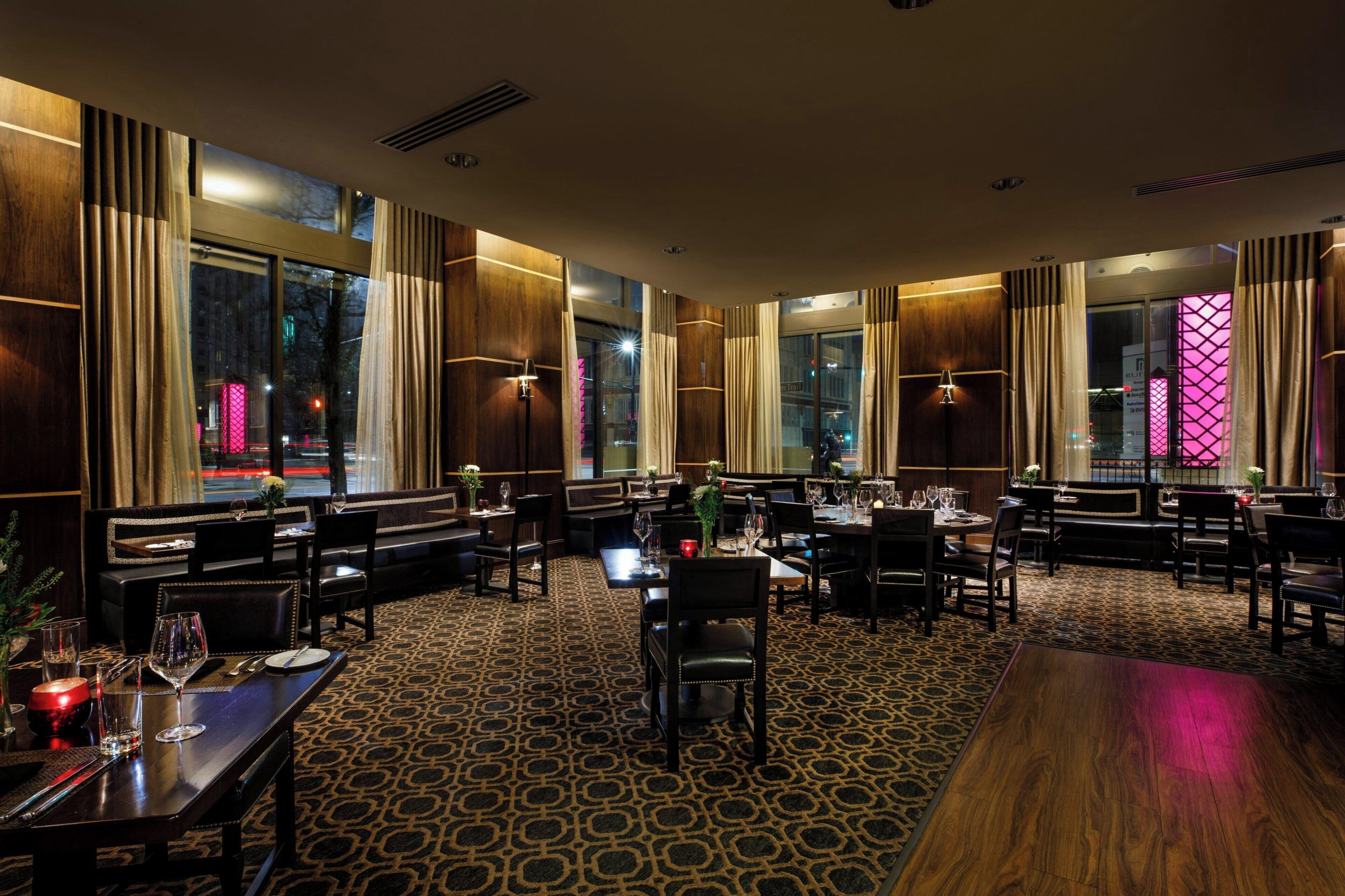 A restaurant with multiple tables and booths, large windows with curtains, and a patterned carpet.