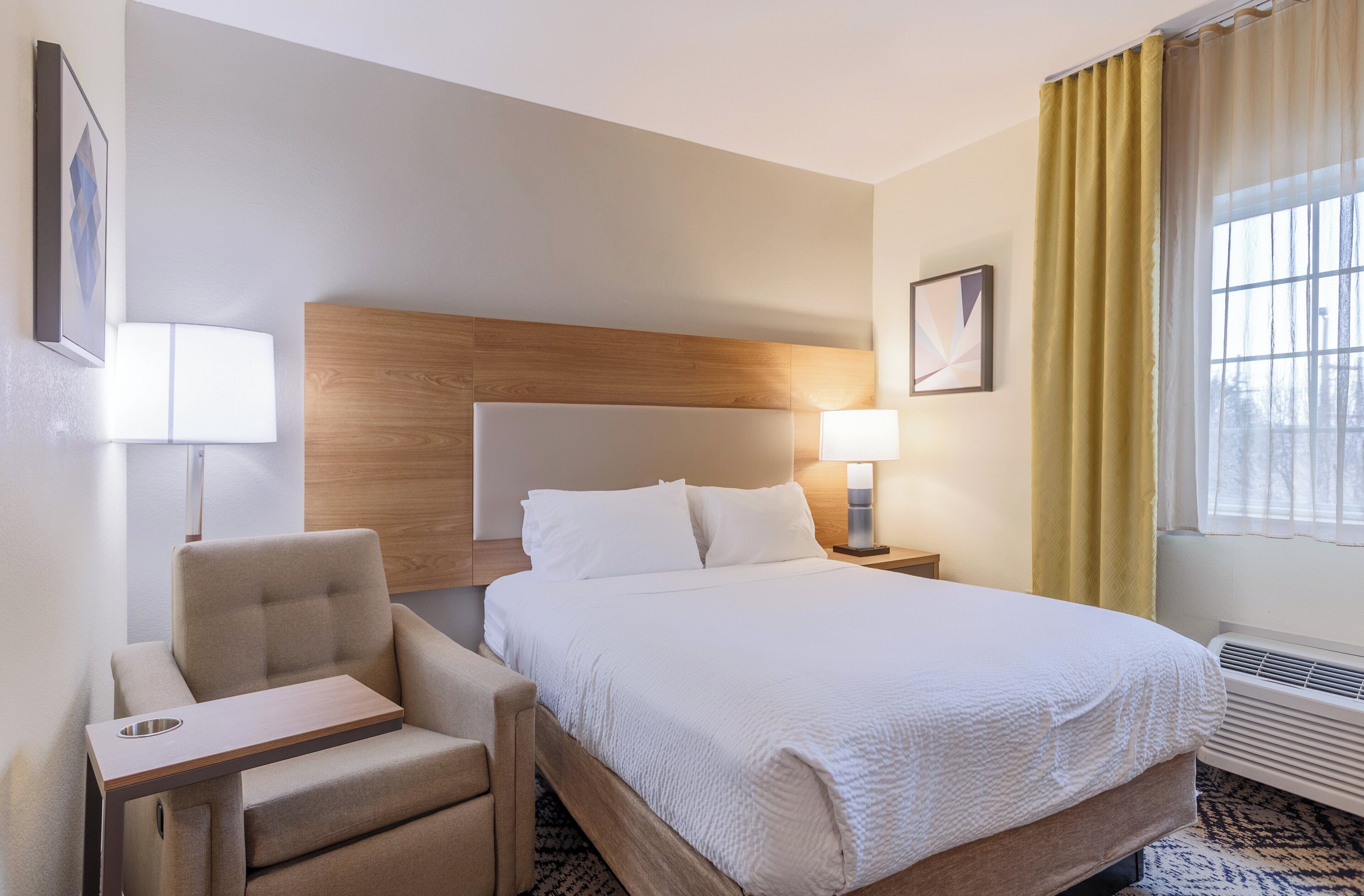 Een hotelkamer met een bed, een stoel, een nachtkastje, een lamp en een raam met gordijnen.