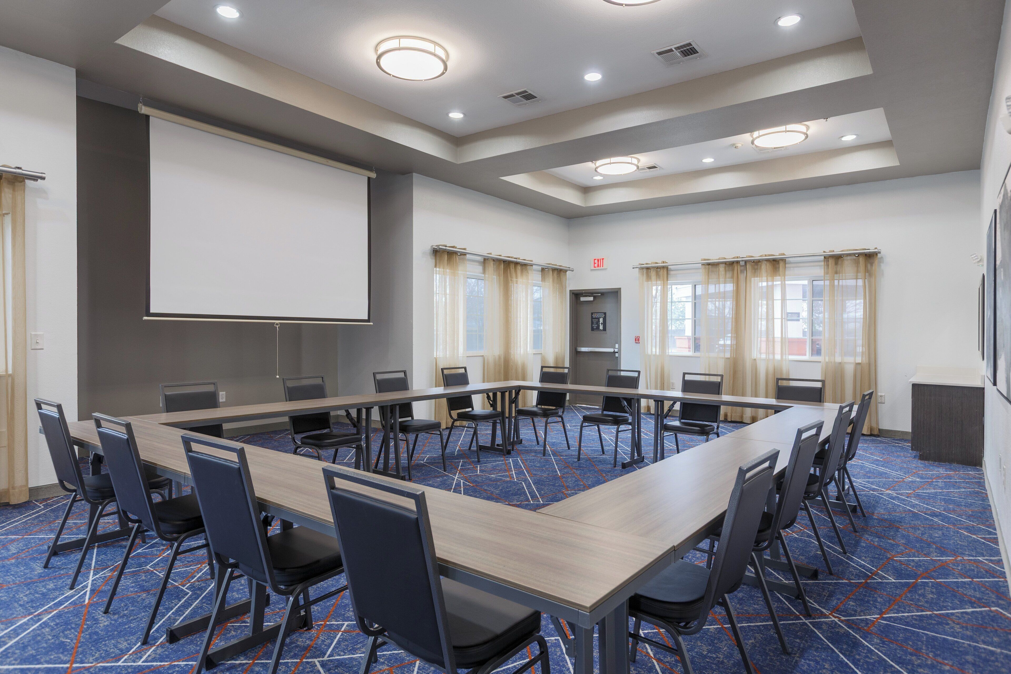 A conference room with a long table, chairs, a projector screen, and windows with curtains.