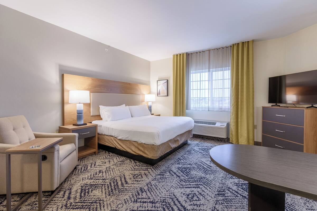 Ein Hotelzimmer mit Bett, Nachttischen, einem Sessel, einem Schreibtisch, einem Fernseher und einem fenster mit Vorhängen.